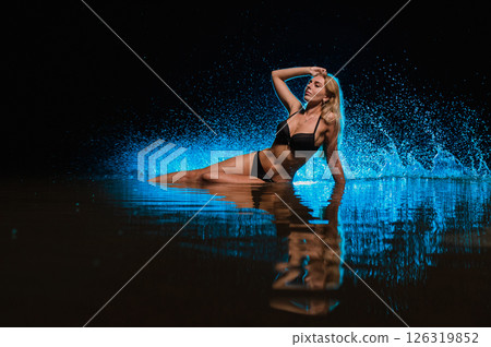 Young slender girl in a swimsuit posing in the water at sunset at the background of blue colored water droplets flying in the air 126319852
