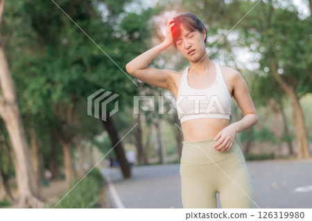 Sport southeast asian female teen jogging running health public park pain during exercise as she stops and touches her head with pained expression in green outdoor setting 126319980