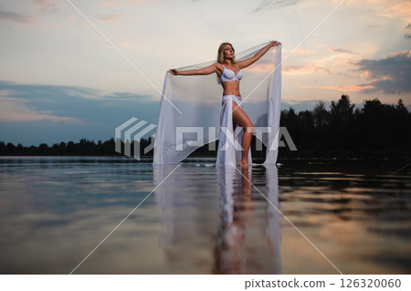 Young slender girl in a swimsuit posing in the water at sunset with big piece of fabric 126320060
