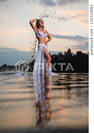 Young slender girl in a swimsuit posing in the water at sunset with big piece of fabric Young slender girl in a swimsuit posing in the water at sunset with big piece of fabric 126320062