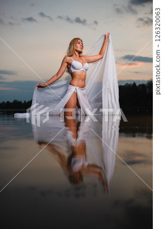 Young slender girl in a swimsuit posing in the water at sunset with big piece of fabric 126320063