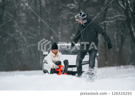 Happy couple are outdoors at winter time. Man takes his wife on a sled. White car behind 126320293