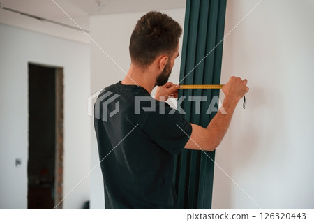 Taking measurements. Plumber worker installing heating radiator in empty room of a newly built house Taking measurements. Plumber worker installing heating radiator in empty room of a newly built house 126320443