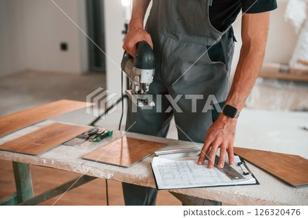 Standing by the table with jigsaw. Man is installing new laminated wooden floor Standing by the table with jigsaw. Man is installing new laminated wooden floor 126320476