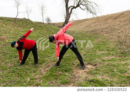 Two individuals engage in stretching exercises on a grassy hillside under a cloudy sky, demonstrating commitment to fitness and wellness in a serene outdoor setting Two individuals engage in stretching exercises on a grassy hillside under a cloudy sky, demonstrating commitment to fitness and wellness in a serene outdoor setting 126320522