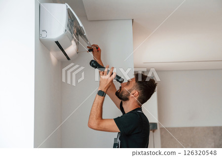 Using screwdriver and flashlight. Man repairs an air conditioner indoors at home Using screwdriver and flashlight. Man repairs an air conditioner indoors at home 126320548