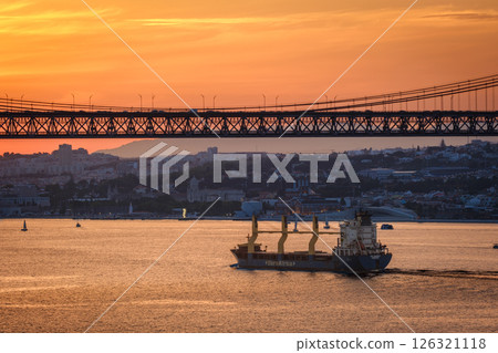 Cargo ship sail on Tagus river under iconic 25 de Abril bridge in silhouette, Lisbon, Portugal at sunset. City skyline, glowing orange sky, dramatic maritime and urban scene, European port, summer 126321118