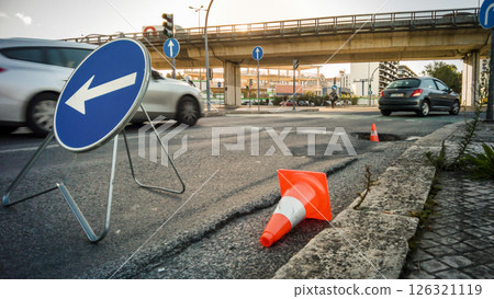 City street with large pothole marked by traffic cones and detour sign, cars passing by and overpass in background. Urban infrastructure issues, road safety, busy traffic hours, street repairs 126321119