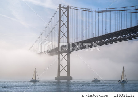 Dramatic view of Lisbon iconic 25 de Abril bridge in morning fog on sunny day, with sailboats and motorboat on tranquil Tagus river, Portugal. Misty atmosphere, serene, mysterious scene, perfect for 126321132