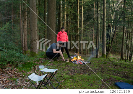 Enchanting camping experience in a serene forest with a warm fire, two friends cooking and enjoying nature's beauty during the golden hour of late afternoon 126321139