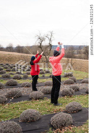 Two individuals engaging in mindful stretching among rows of lavender in a serene countryside setting during a cloudy winter day, promoting wellness and connection with nature Two individuals engaging in mindful stretching among rows of lavender in a serene countryside setting during a cloudy winter day, promoting wellness and connection with nature 126321145