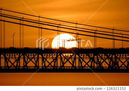 Close view of sun setting directly behind 25 de Abril bridge in Lisbon, Portugal, warm orange sky with some clouds, cars and cables in silhouette against glowing sun, striking urban sunset scene 126321152