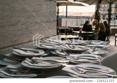 Plates neatly arranged with forks, knives, and napkins on stone countertop at outdoor restaurant in selective focus. Blurred background with European city square at sunset. Relaxed dining, people 126321155