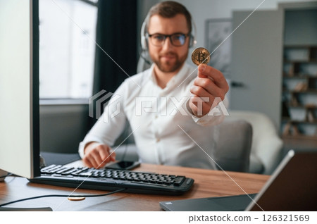 Holding bitcoin in the hand. Man in formal clothes is working in the modern office. Using computer Holding bitcoin in the hand. Man in formal clothes is working in the modern office. Using computer 126321569