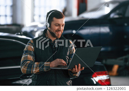Smiling, standing, holding laptop. In headphones. Man in uniform is working in the auto salon 126322001