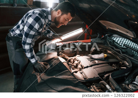 Under the hood. Holding lighting equipment and fixing the issues of the car. Man in uniform is working in the auto salon 126322002