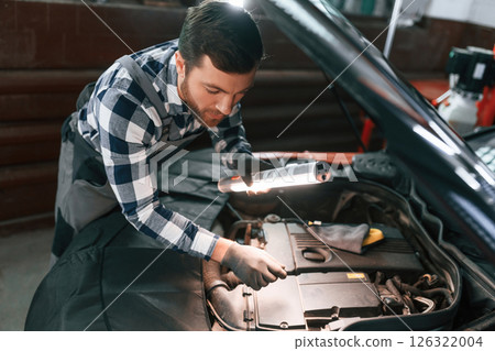 Under the hood. Holding lighting equipment and fixing the issues of the car. Man in uniform is working in the auto salon 126322004