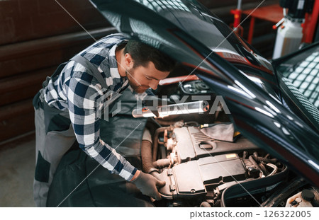 Under the hood. Holding lighting equipment and fixing the issues of the car. Man in uniform is working in the auto salon 126322005