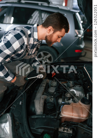 Mechanic is using a tool to repair the car under the hood. Man in uniform is working in the auto salon 126322051