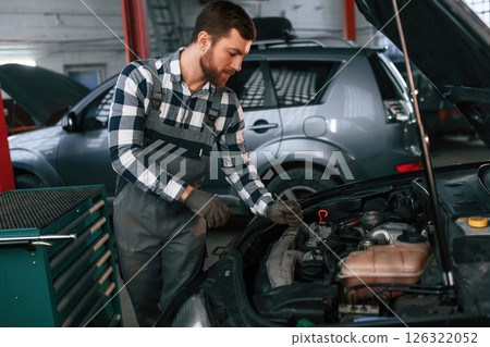Mechanic is using a tool to repair the car under the hood. Man in uniform is working in the auto salon 126322052