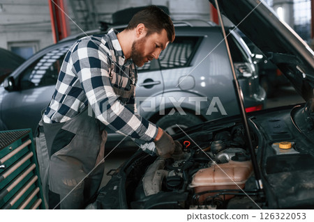 Mechanic is using a tool to repair the car under the hood. Man in uniform is working in the auto salon 126322053