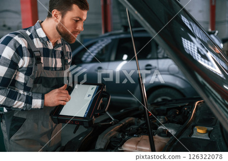 Standing near the car with hood opened. Holding tablet. Man in uniform is working in the auto salon Standing near the car with hood opened. Holding tablet. Man in uniform is working in the auto salon 126322078