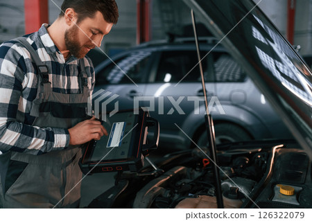 Standing near the car with hood opened. Holding tablet. Man in uniform is working in the auto salon Standing near the car with hood opened. Holding tablet. Man in uniform is working in the auto salon 126322079