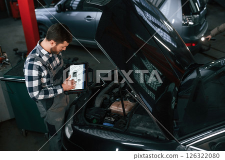 Standing near the car with hood opened. Holding tablet. Man in uniform is working in the auto salon 126322080