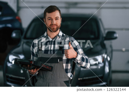 Showing gesture and smiling. Holding tablet. Man in uniform is working in the auto salon Showing gesture and smiling. Holding tablet. Man in uniform is working in the auto salon 126322144