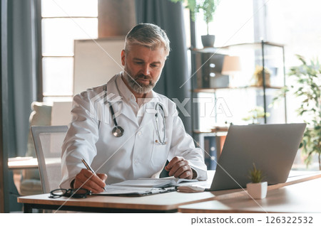 Sitting by table with laptop, writing data into document. Senior doctor in white coat is at work indoors 126322532