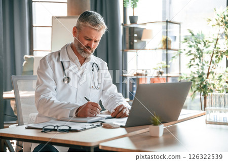 Sitting by table with laptop, writing data into document. Senior doctor in white coat is at work indoors Sitting by table with laptop, writing data into document. Senior doctor in white coat is at work indoors 126322539