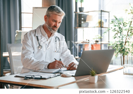 Sitting by table with laptop, writing data into document. Senior doctor in white coat is at work indoors Sitting by table with laptop, writing data into document. Senior doctor in white coat is at work indoors 126322540