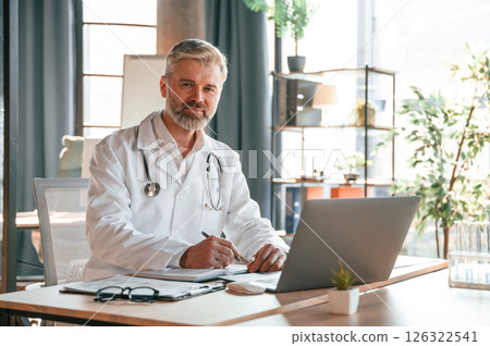 Sitting by table with laptop, writing data into document. Senior doctor in white coat is at work indoors Sitting by table with laptop, writing data into document. Senior doctor in white coat is at work indoors 126322541