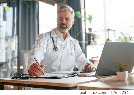 Writing information into document. Senior doctor in white coat is at work indoors 126322544