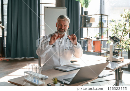 Sitting and smiling. Senior doctor in white coat is at work indoors 126322555