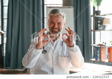 Sitting and smiling. Senior doctor in white coat is at work indoors 126322557