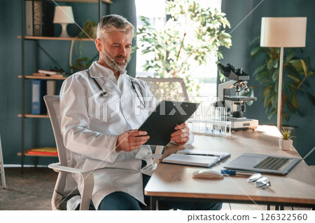 Reading the document from a notepad. Senior doctor in white coat is at work indoors Reading the document from a notepad. Senior doctor in white coat is at work indoors 126322560