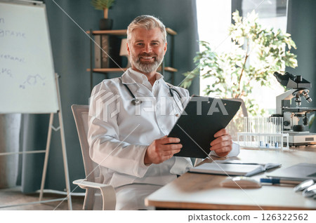 Sitting and smiling. Senior doctor in white coat is at work indoors 126322562