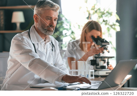Using microscope. Two clinic workers are indoors together. Senior man with young woman 126322720