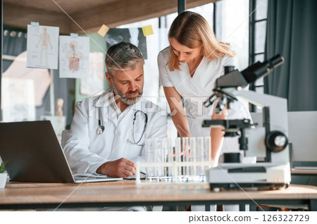 Laptop, test tubes, microscope. Two clinic workers are indoors together. Senior man with young woman 126322729