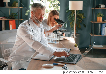 Laptop and microscope are on the table. Two clinic workers are indoors together. Senior man with young woman 126322754