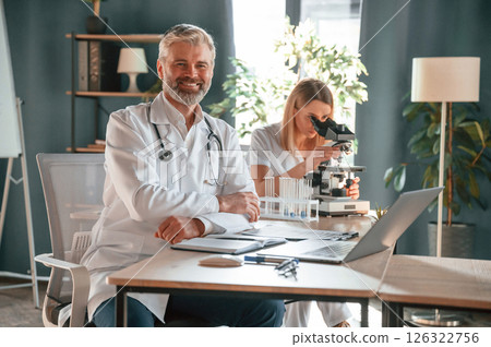 Man is smiling, woman is using microscope. Two clinic workers are indoors together Man is smiling, woman is using microscope. Two clinic workers are indoors together 126322756