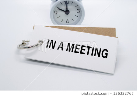 In A Meeting text on white notepad with alarm clock on white background. Away from office concept In A Meeting text on white notepad with alarm clock on white background. Away from office concept 126322765