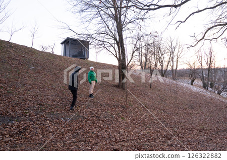 Two individuals exploring a leaf covered hillside during early evening in a tranquil landscape with bare trees and a small shelter in the background 126322882