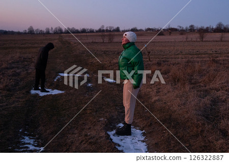 Two figures stand in a rural landscape at dusk, one dressed warmly in green while the other quietly bows, capturing moments of solitude and reflection in nature Two figures stand in a rural landscape at dusk, one dressed warmly in green while the other quietly bows, capturing moments of solitude and reflection in nature 126322887