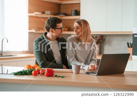 Sitting, using laptop. Young couple is on the kitchen together Sitting, using laptop. Young couple is on the kitchen together 126322990