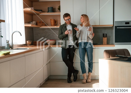 Standing, talking, holding cups with drink. Young couple is on the kitchen together Standing, talking, holding cups with drink. Young couple is on the kitchen together 126323004
