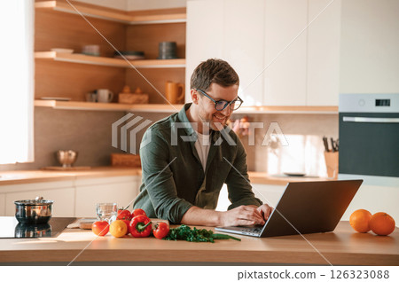 Sitting by table and using laptop. Handsome man is on the kitchen at daytime 126323088