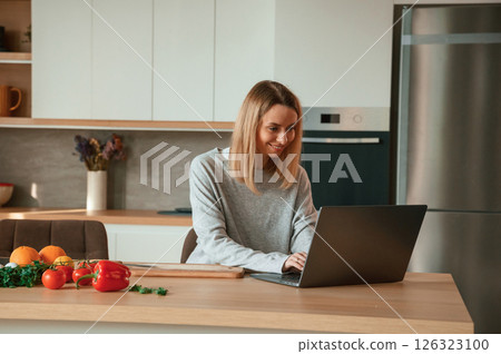 Sitting by laptop. Vegetables are on the kitchen. Beautiful young woman is on the kitchen 126323100