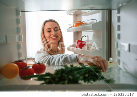 Vegetables is in the fridge. Beautiful young woman is on the kitchen 126323117